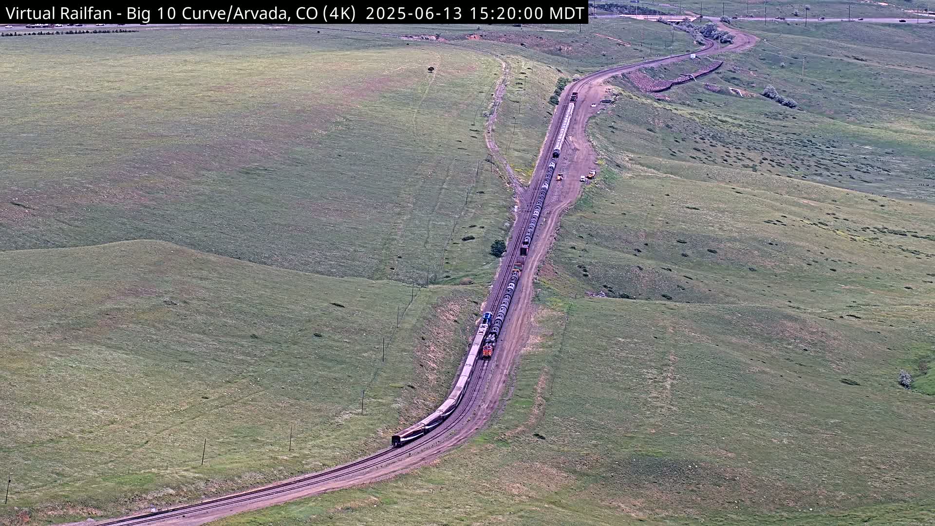 A long freight train curves around a hillside on a railway track through grassy terrain under a clear sky.