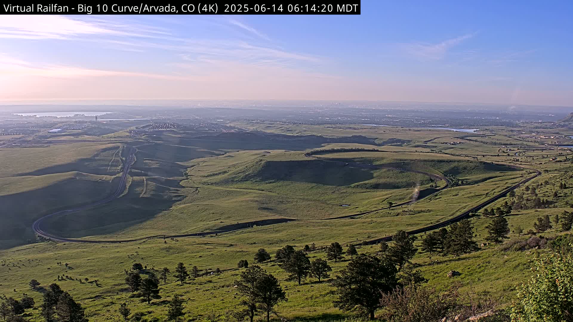 A winding road curves through a grassy landscape under a clear, bright sky, with a distant city visible on the horizon.