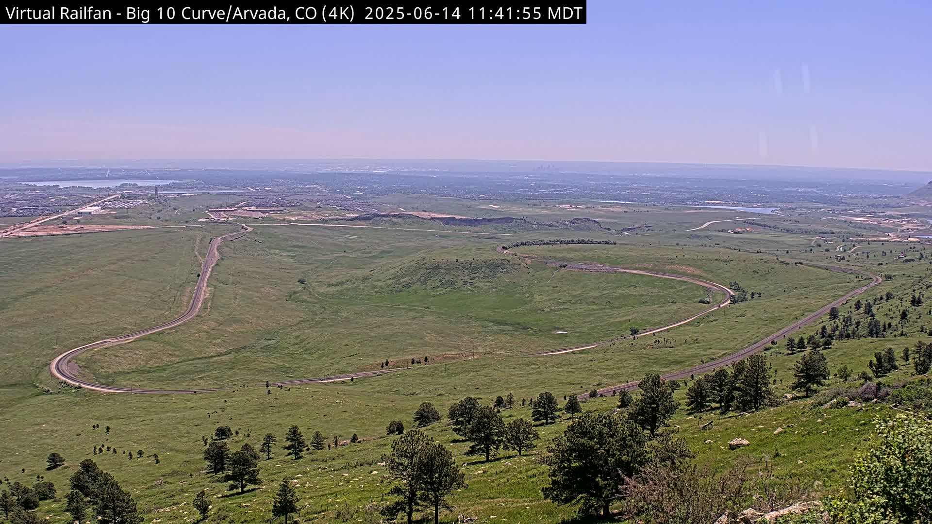 A winding road curves through a grassy landscape under a clear, bright sky, with a distant city visible on the horizon.