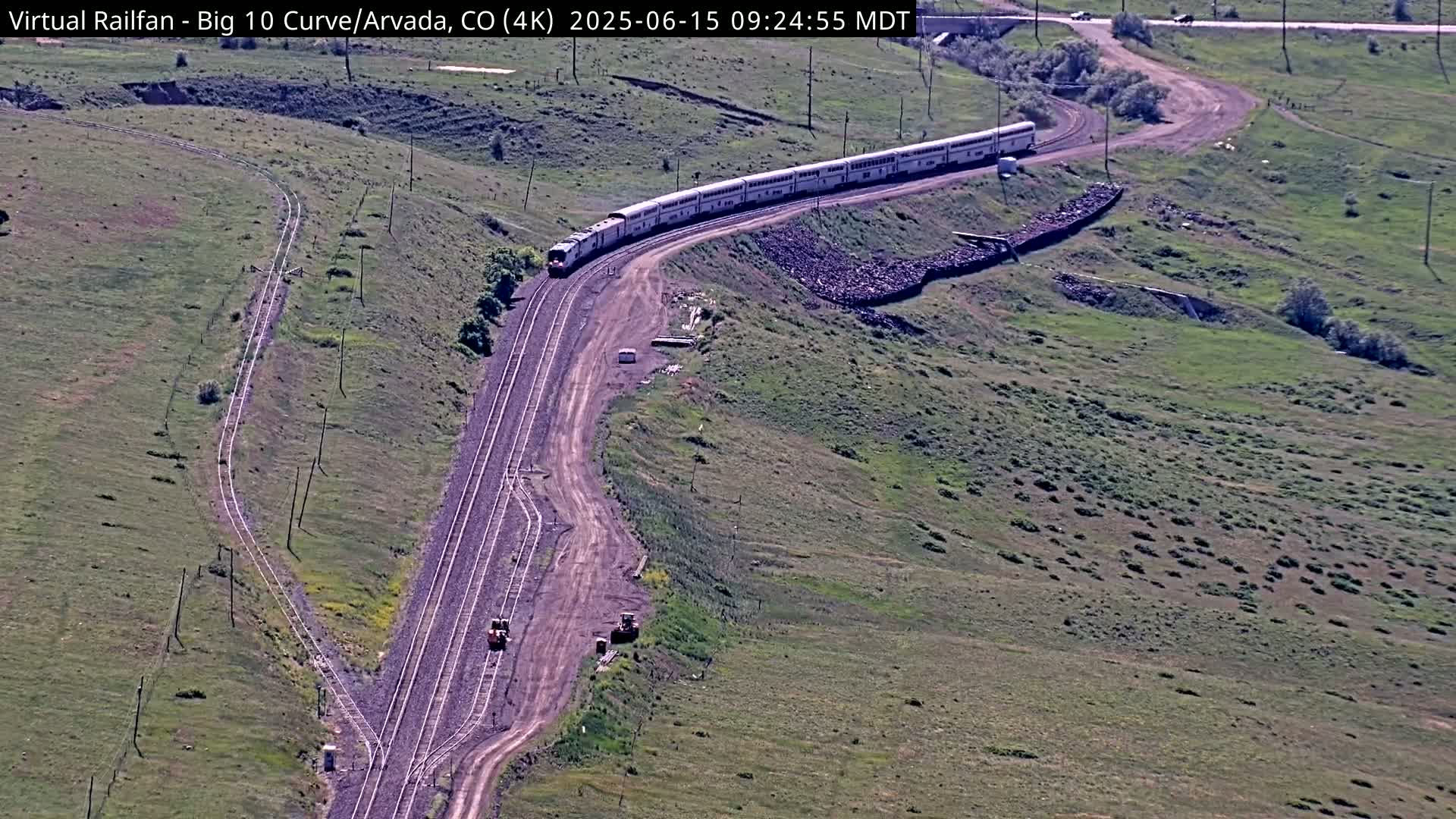 A long passenger train rounds a curve on a hillside, with construction equipment visible near the tracks, under sunny conditions.