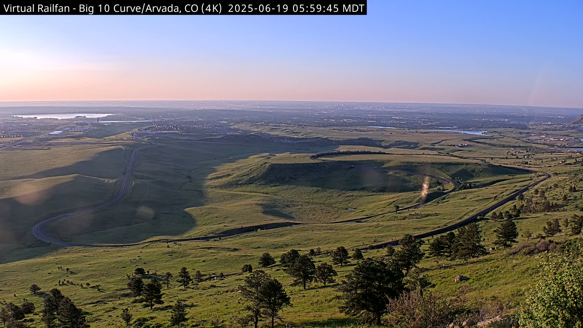 A winding road curves through a vast, grassy landscape under a clear, sunrise sky, with a distant city visible on the horizon.