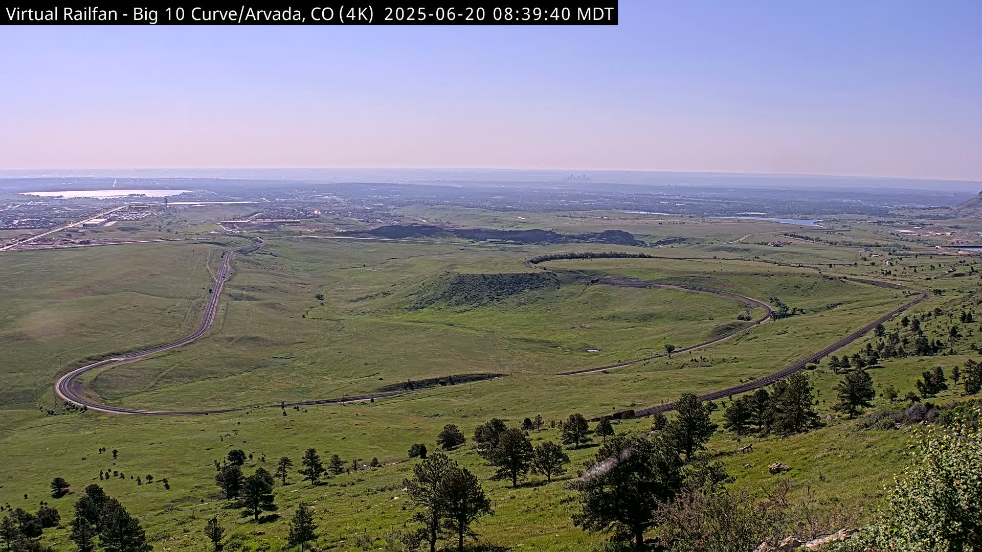 A high-angle, long shot on a clear day shows a winding road curving through a grassy landscape, with a distant city skyline visible on the horizon.
