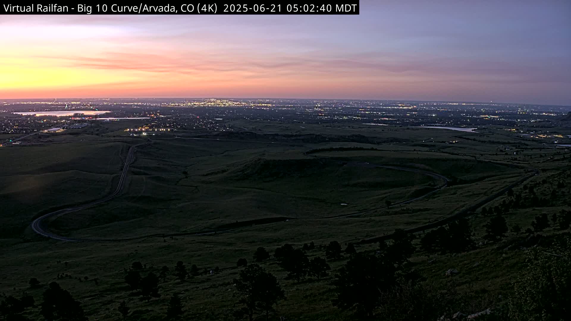 A winding road curves through a dark green landscape at twilight, overlooking a distant city illuminated by lights under a dusky pink and purple sky.