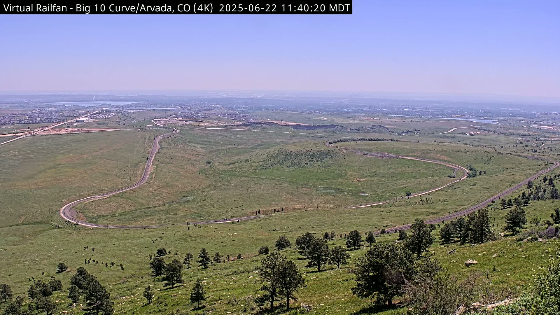 A winding road curves through a grassy landscape under a clear, hazy sky, with a distant view of a city and bodies of water.