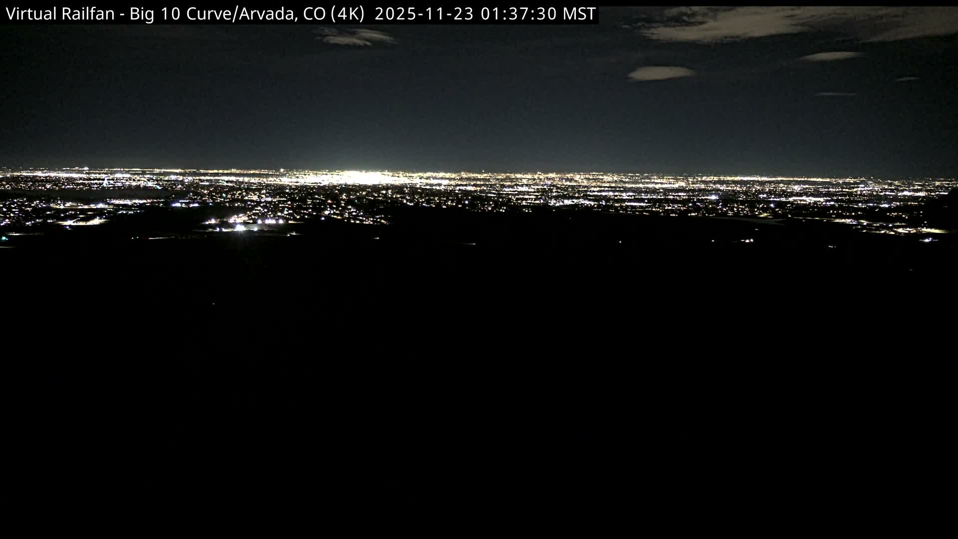 A vast cityscape sparkles with countless lights under a dark, partly cloudy night sky, viewed from an elevated perspective.