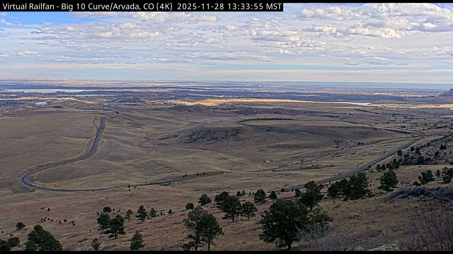Under a partly cloudy sky, a winding railroad track traverses dry, golden-brown hills dotted with sparse trees, leading towards a distant urban landscape with visible bodies of water.