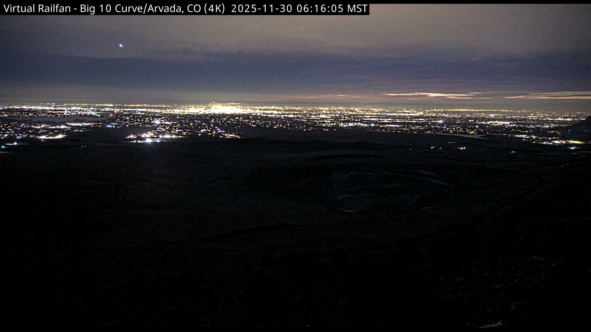 Under a partly cloudy sky, a winding railroad track traverses dry, golden-brown hills dotted with sparse trees, leading towards a distant urban landscape with visible bodies of water.