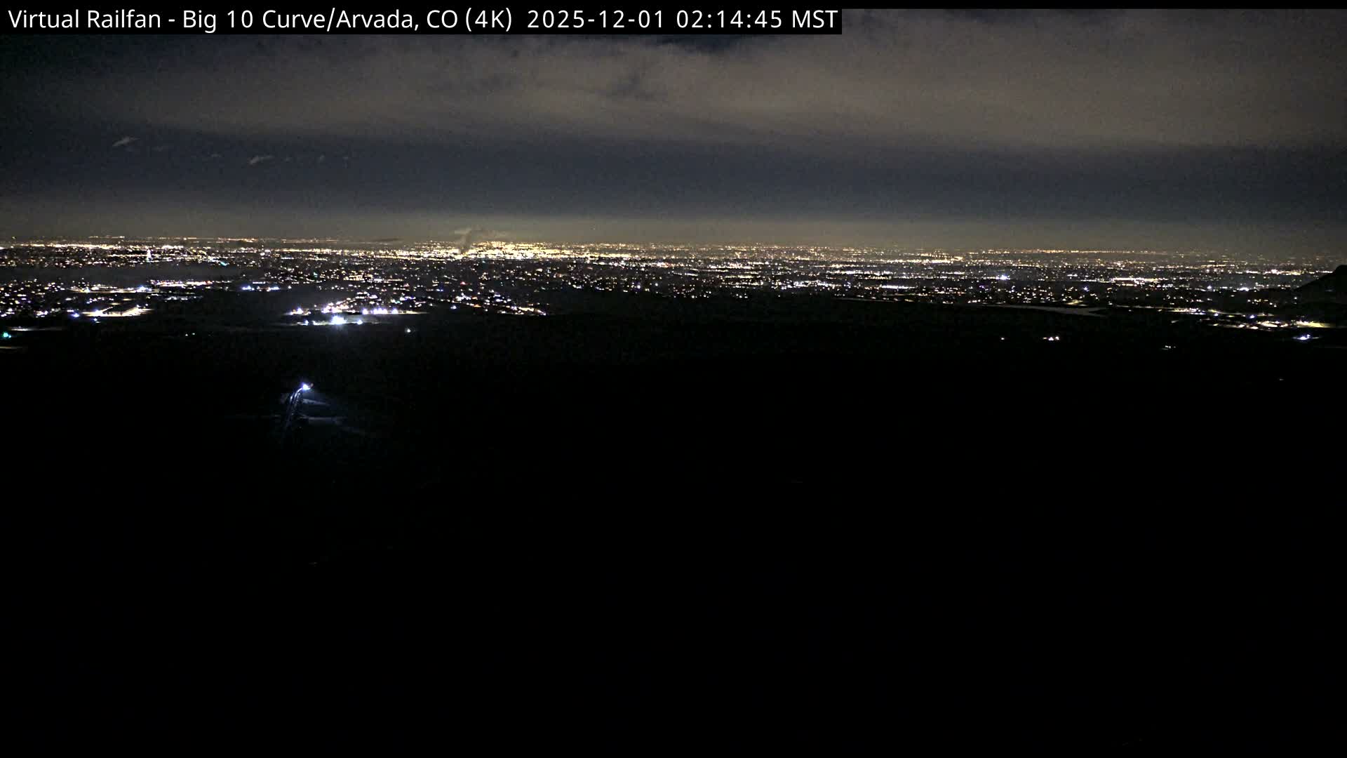 A panoramic nighttime view reveals a vast expanse of city lights stretching into the distance under a dark, cloudy sky.