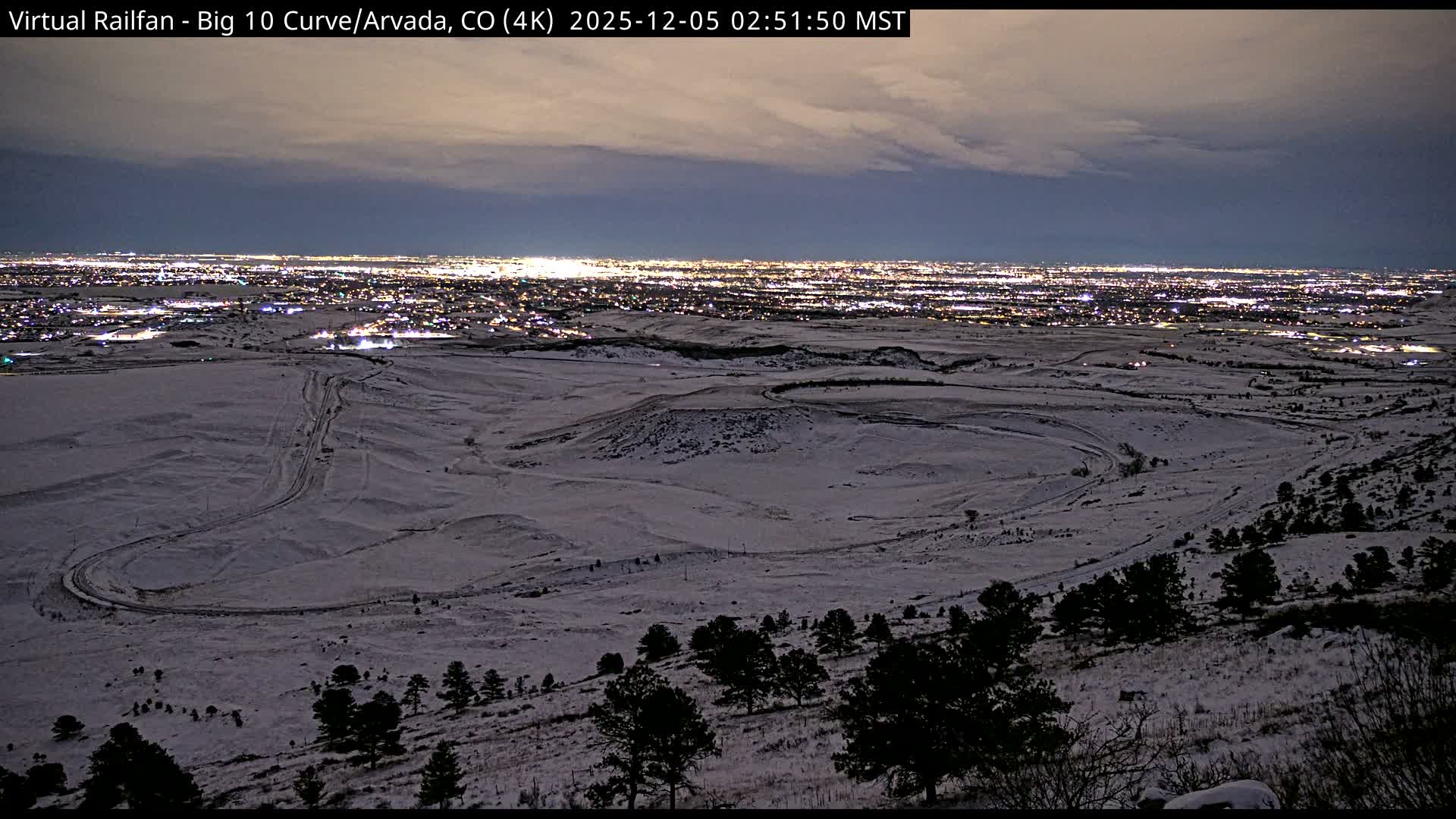 A panoramic view reveals a snow-covered landscape with a prominent curving path in the foreground, leading towards a vast, brightly illuminated city skyline under an overcast night sky.
