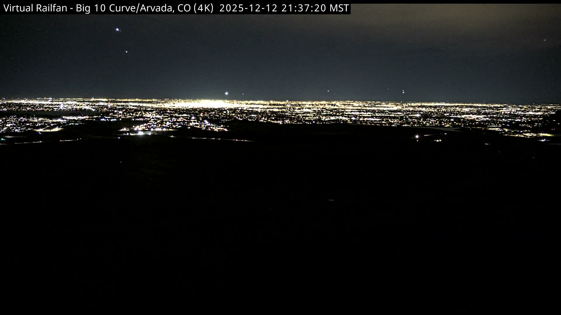 A vast, brightly lit cityscape sprawls across the distant horizon under a dark, clear night sky, viewed from an elevated and unlit foreground.