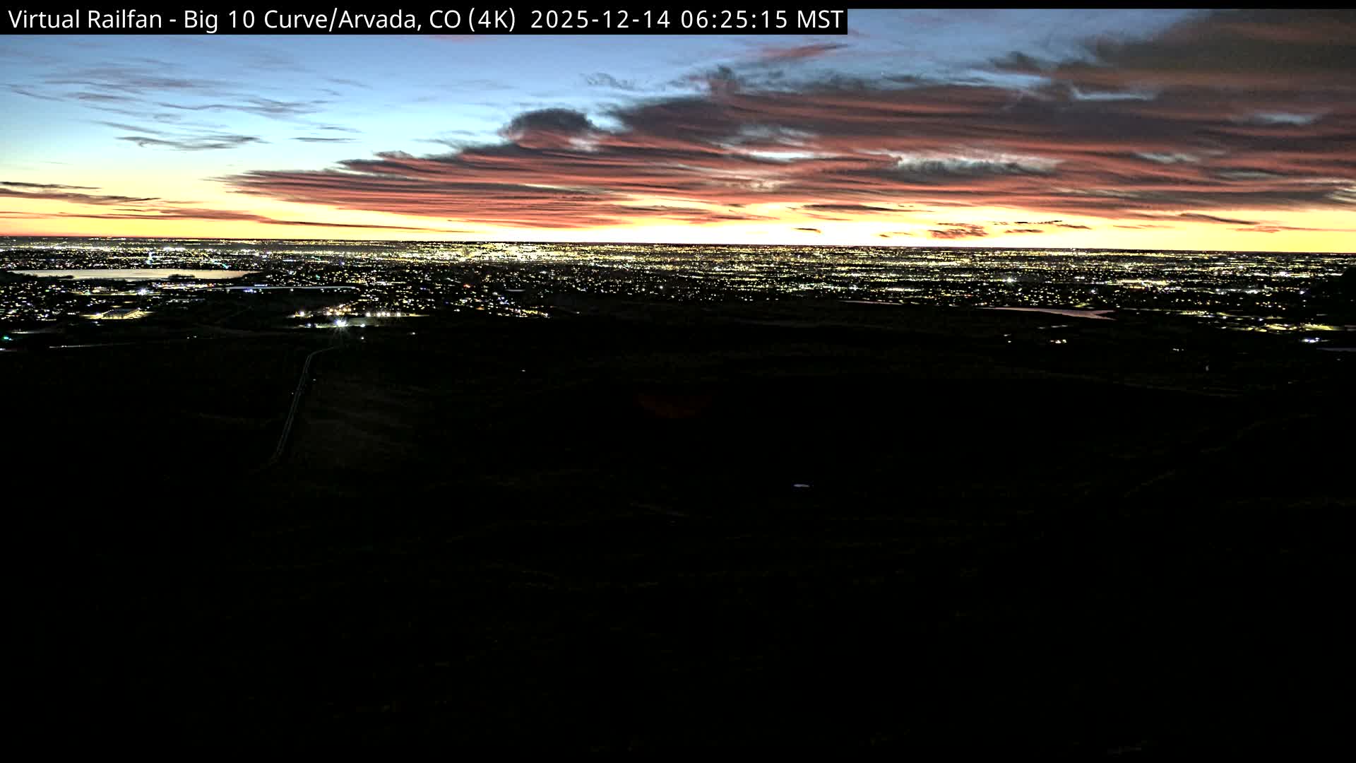 A vast, brightly lit cityscape sprawls across the distant horizon under a dark, clear night sky, viewed from an elevated and unlit foreground.