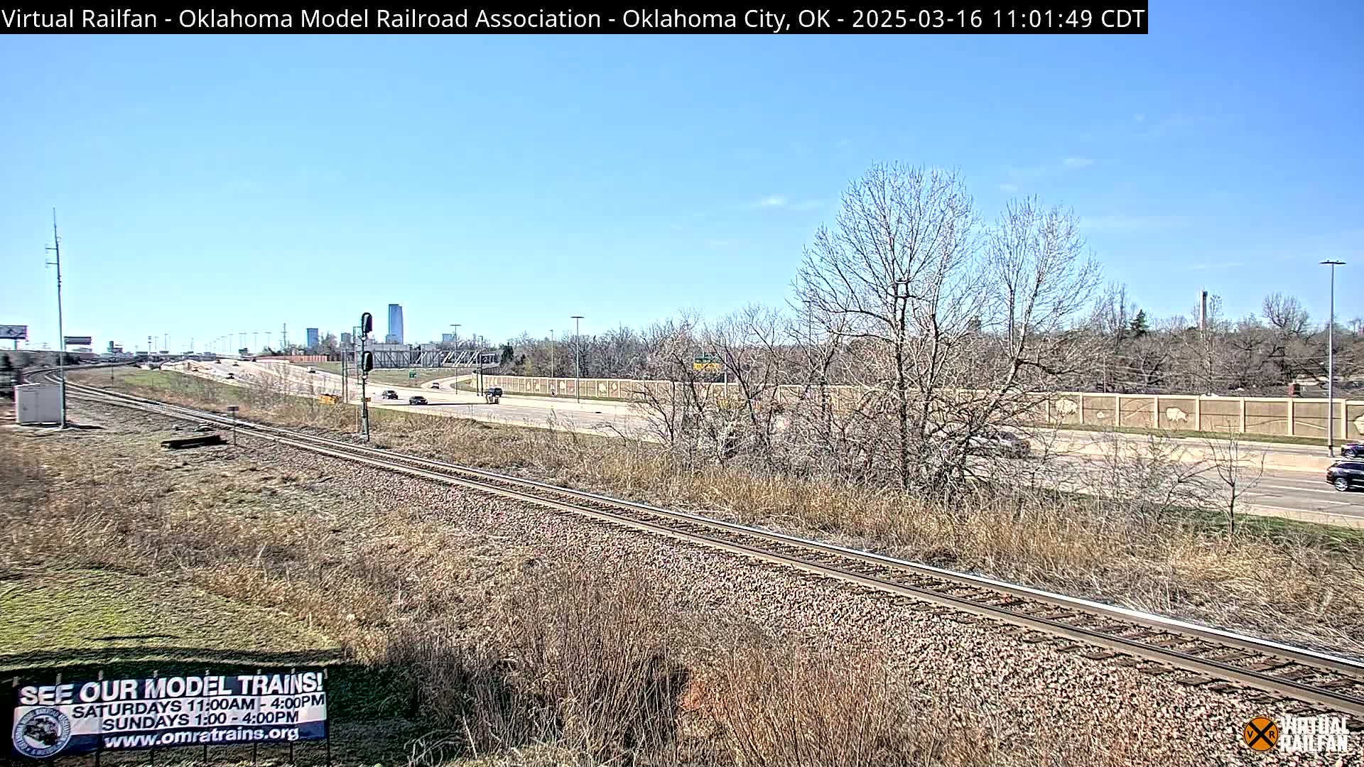 A mostly sunny day reveals a railway track beside a highway, with sparse, dry vegetation and leafless trees surrounding them, and a distant cityscape visible on the horizon.