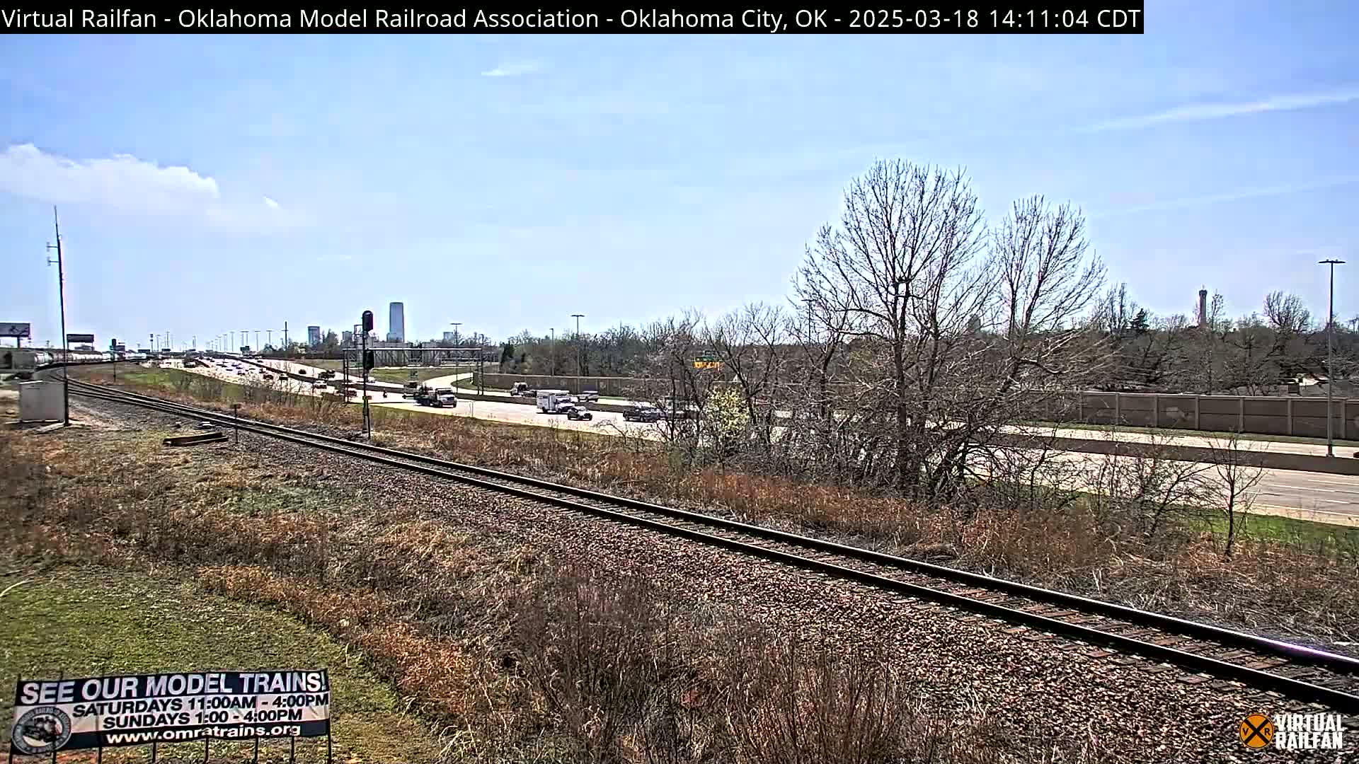 A mostly sunny day shows a highway and railroad tracks running parallel, with sparse, dry-looking vegetation between them, and a cityscape visible in the distance.