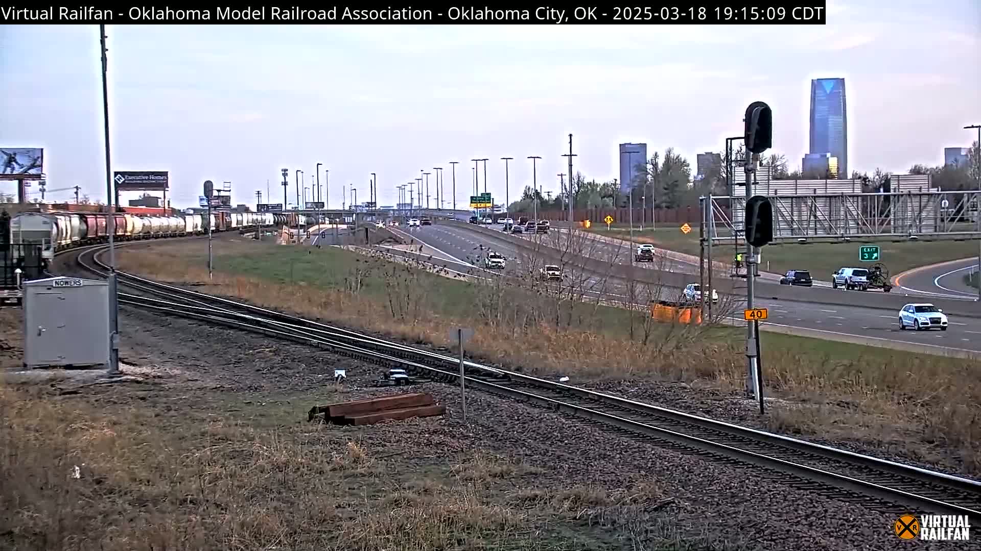A long freight train curves onto a set of tracks beside a highway with sparse traffic under a cloudy sky.