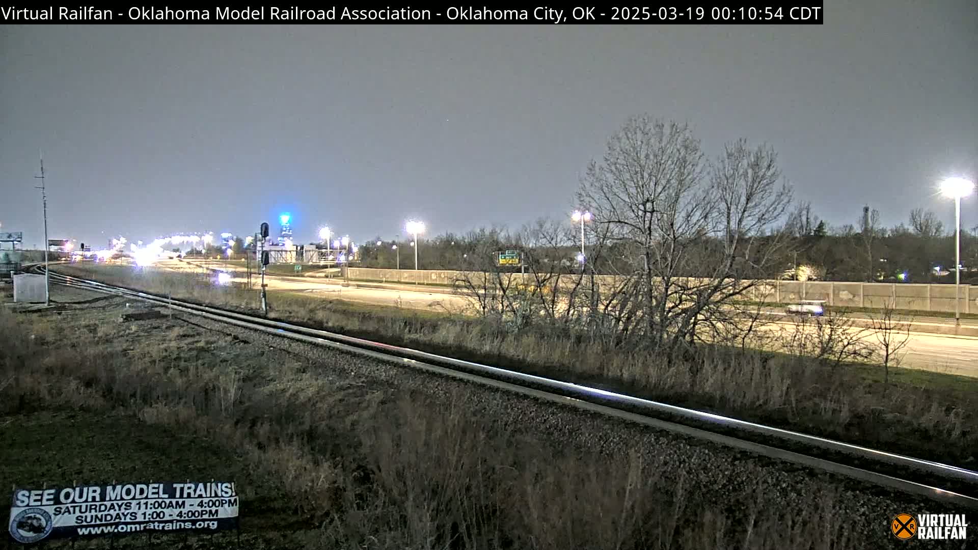 A nighttime view of railroad tracks next to a highway with sparse traffic under a clear, dusky sky.