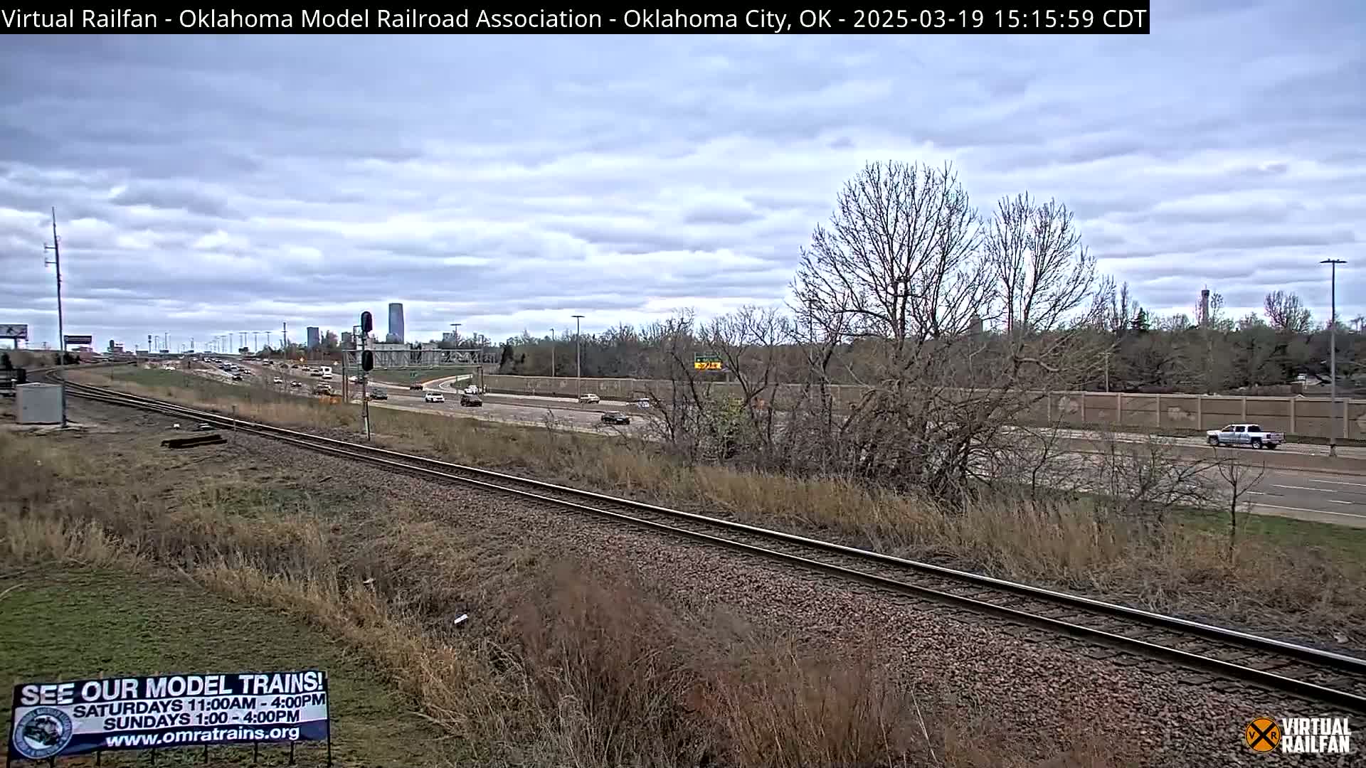 A mostly cloudy day reveals a view of a highway and railway tracks, with sparse, dry vegetation alongside.