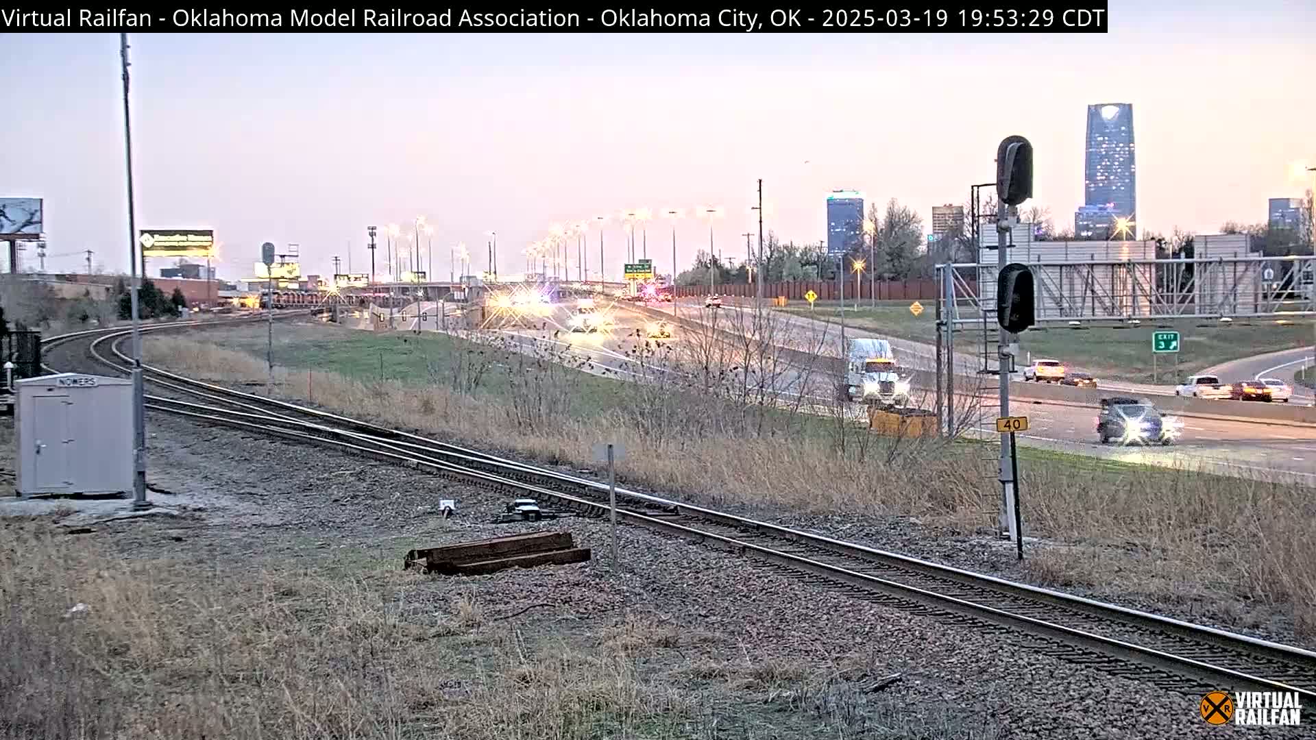 A twilight view shows train tracks next to a highway with several cars and trucks driving under a mostly clear sky.