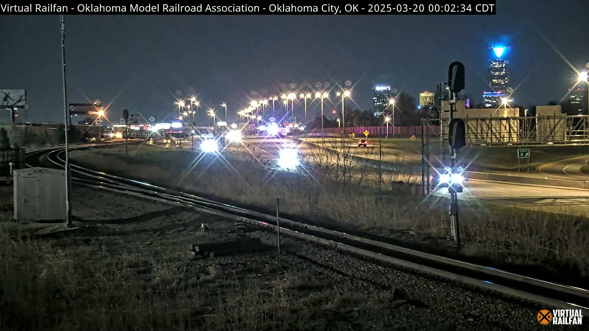 Nighttime view of a highway and train tracks intersecting, with vehicles driving on the highway under illuminated streetlights and a tall building with blue lighting visible in the distance.