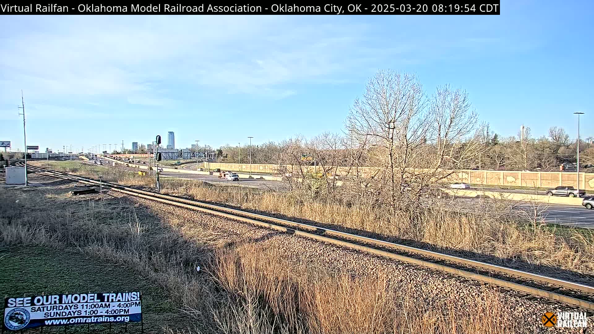 A sunny day shows railroad tracks next to a highway with sparse, dry vegetation and a few cars visible in the distance.