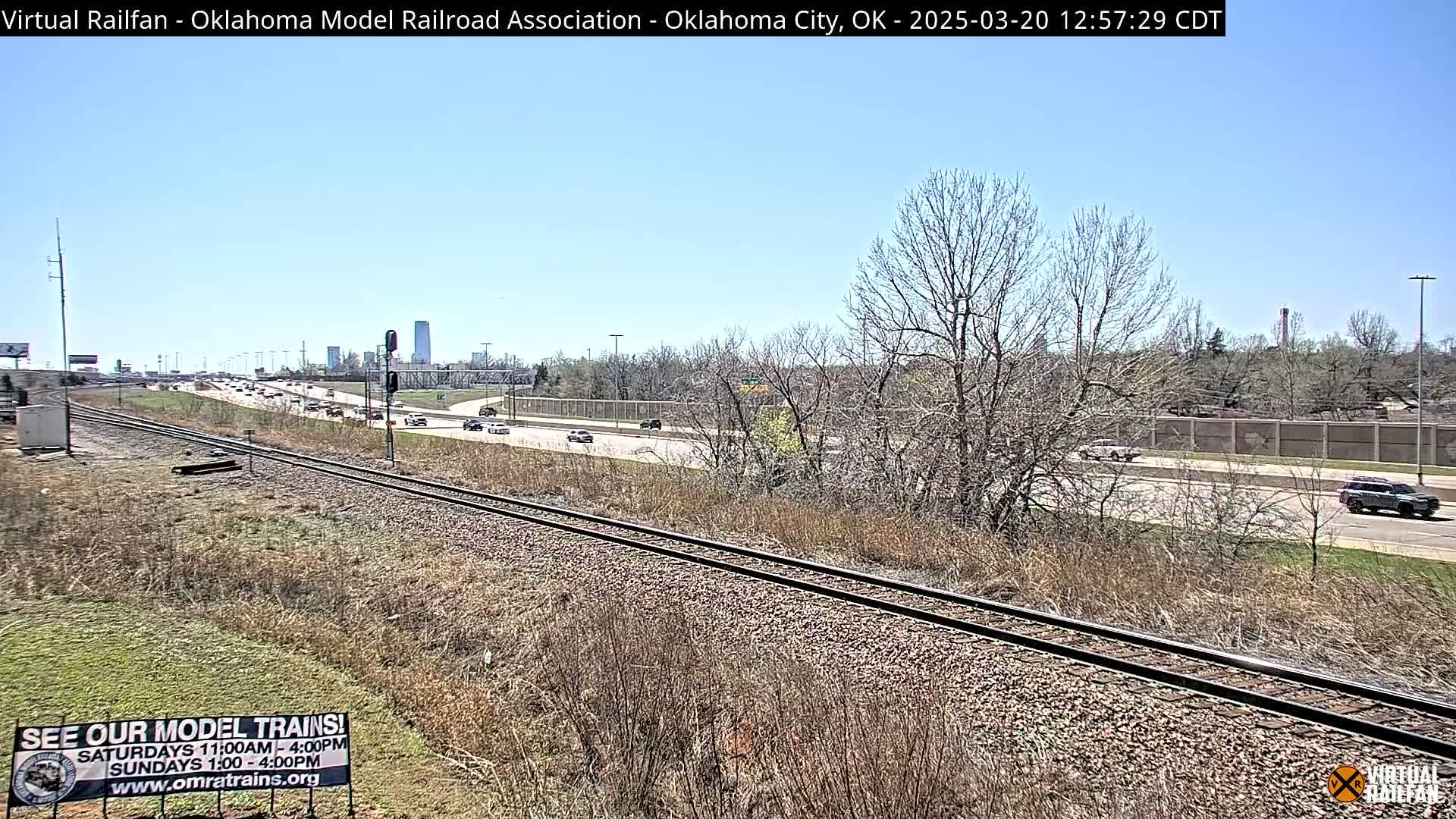 A mostly sunny day shows a view of train tracks next to a highway with sparse, dry vegetation and leafless trees, with a city skyline visible in the distance.
