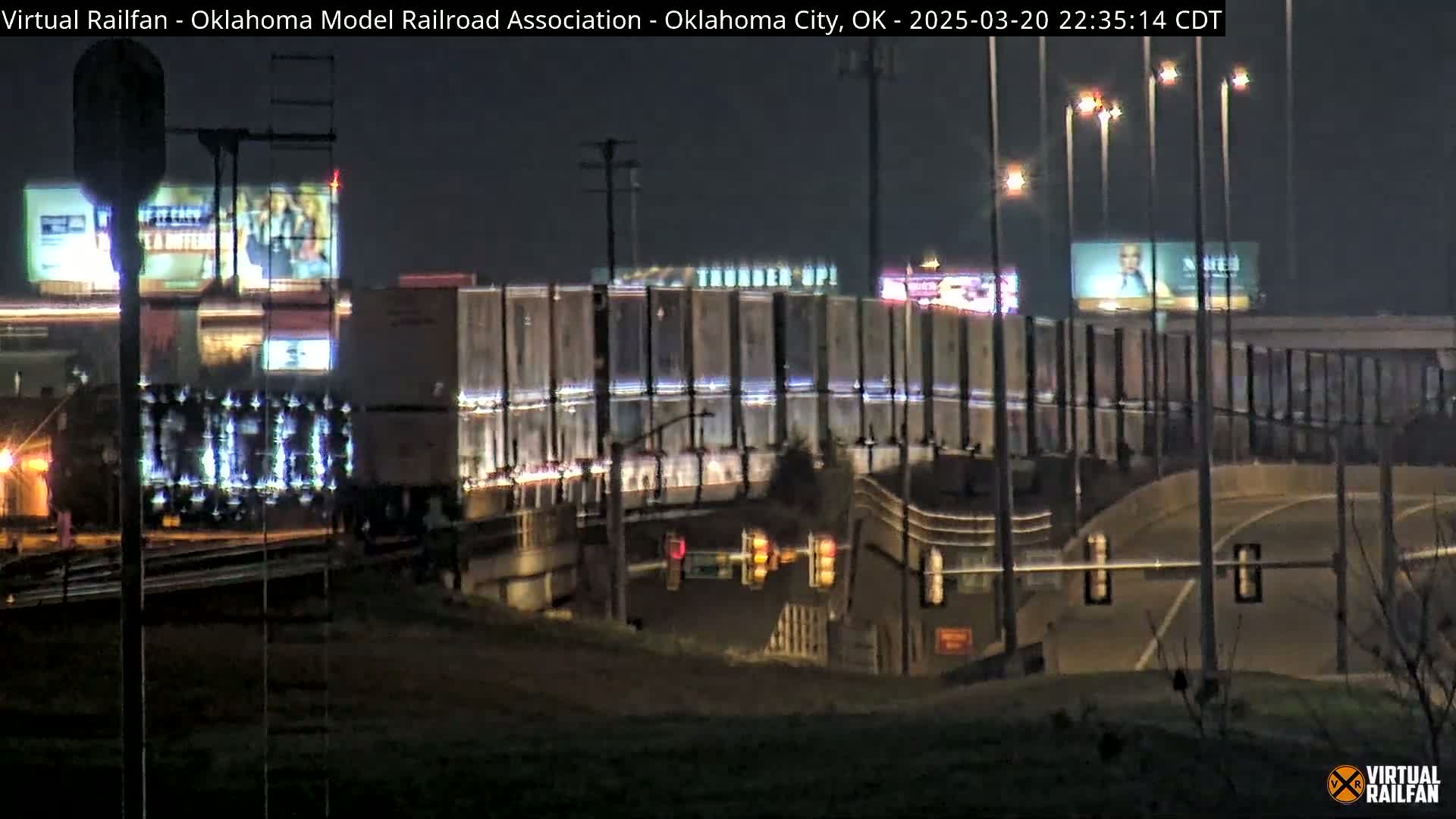 At night, a train passes under a highway overpass with illuminated billboards and streetlights.