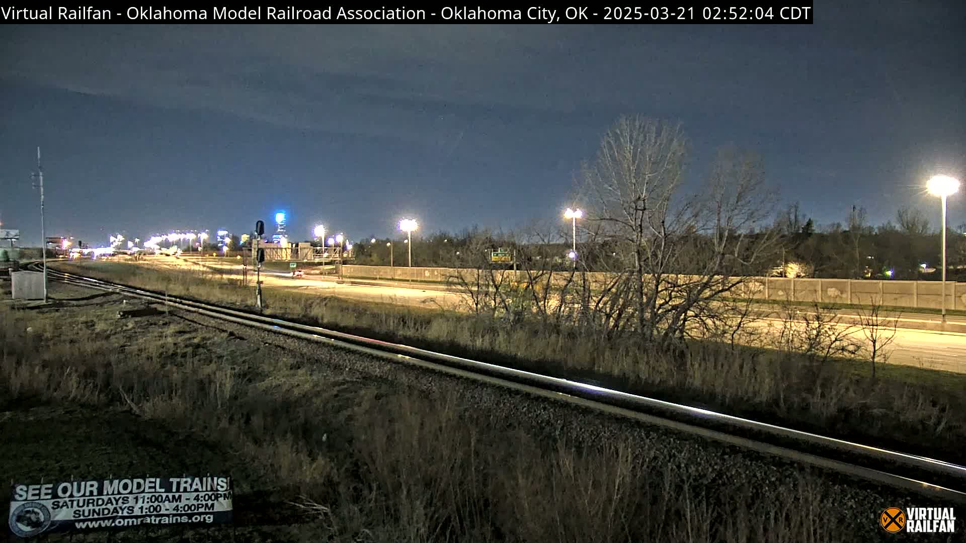 A nighttime view of railroad tracks beside a highway, with sparsely leafed trees and lightly snow-covered ground.