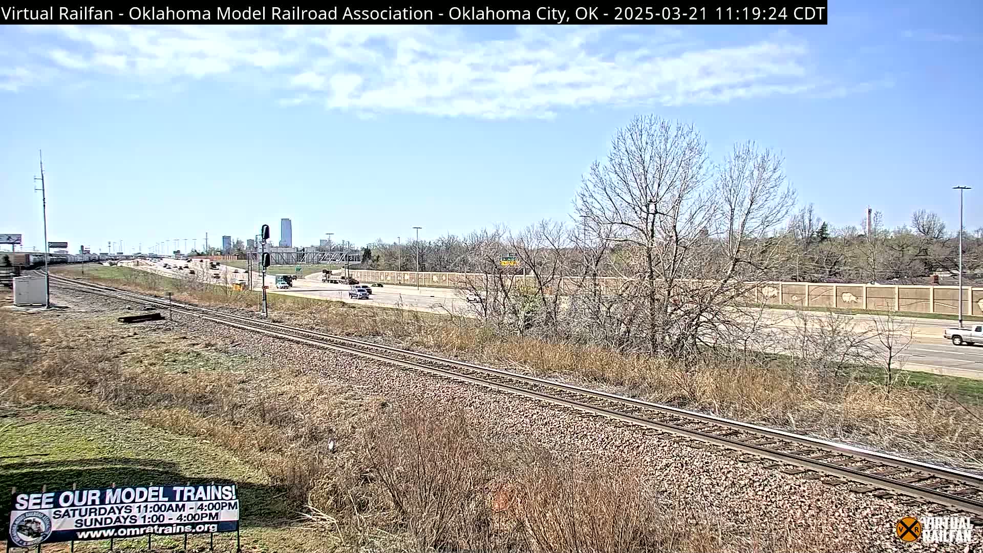 A partly cloudy day reveals a train track alongside a highway with sparse, dry vegetation, a few cars on the road, and a distant cityscape.