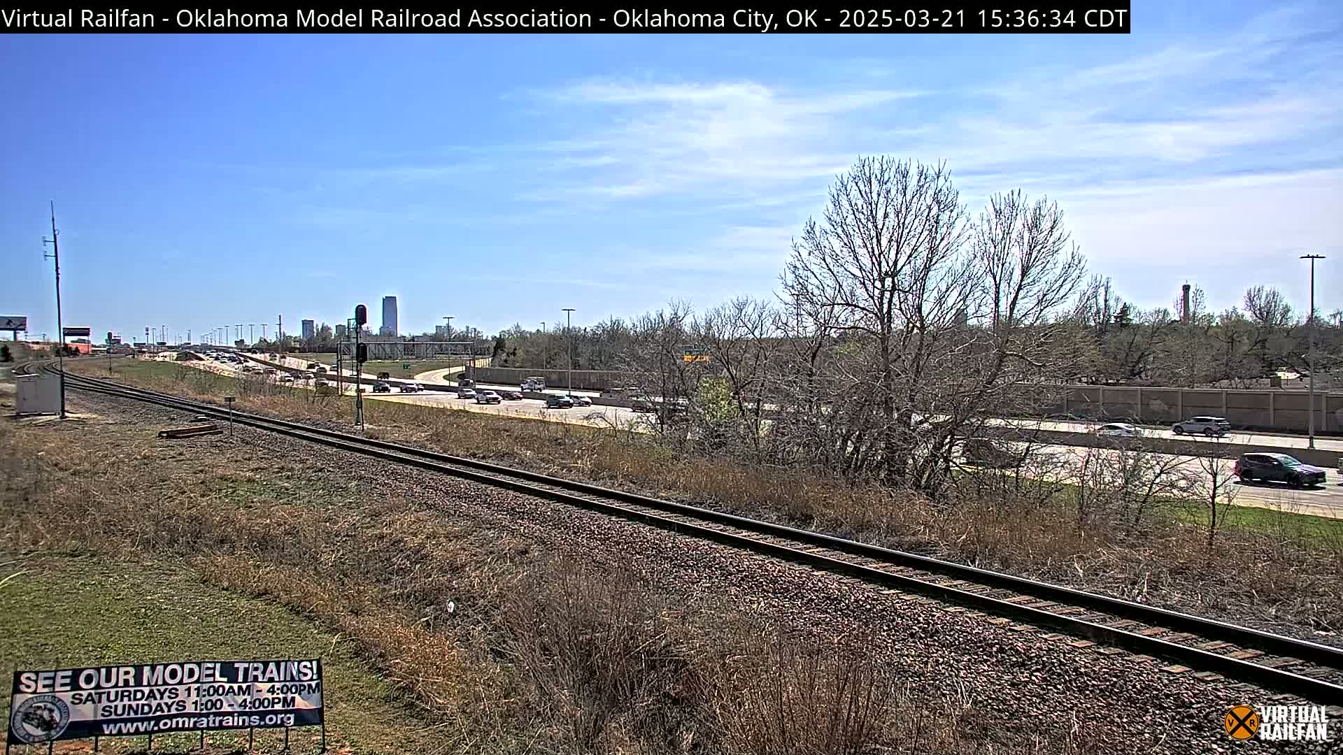 A mostly sunny day shows a highway with moderate traffic alongside a set of train tracks running parallel, with leafless trees and dry, brown grass in the foreground and a cityscape visible in the distance.