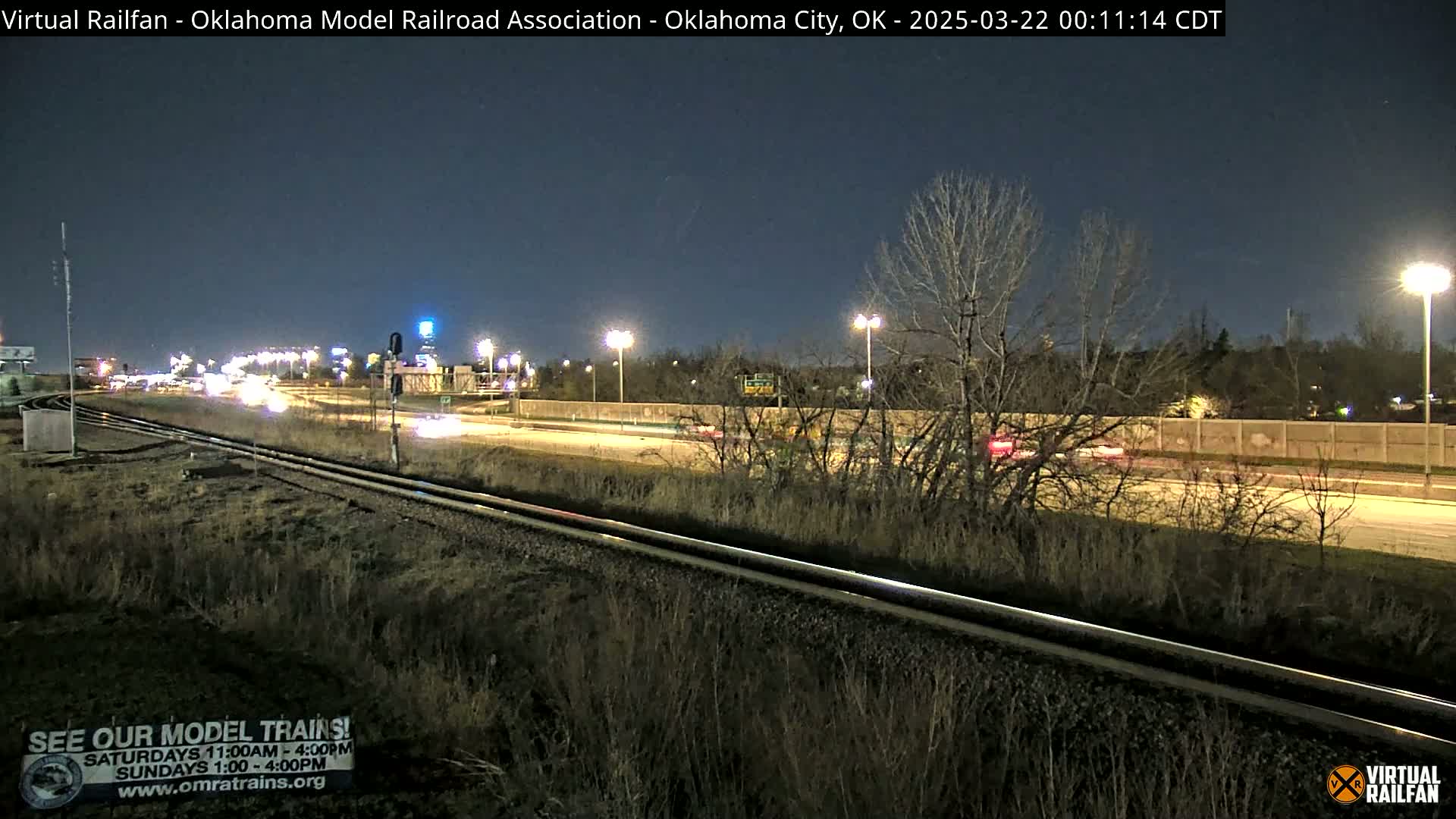 At night, a highway with cars driving on it is adjacent to a set of train tracks, with sparse vegetation and trees between them, under a clear dark sky.