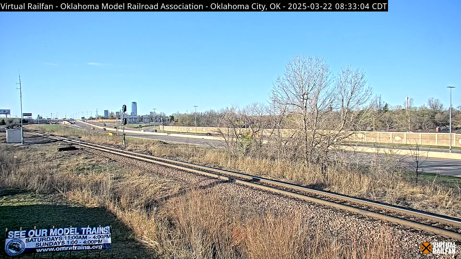 A sunny day reveals railroad tracks next to a highway, with a wall separating them, and bare trees and dry grass in the foreground.