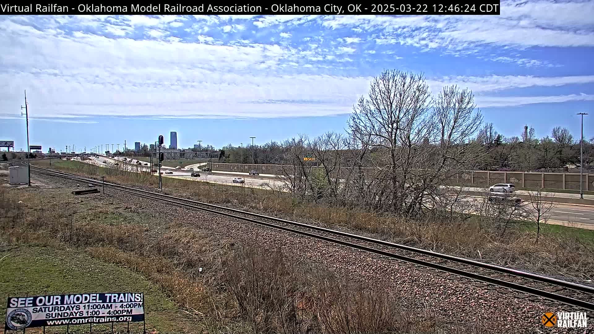 A mostly sunny day reveals a highway with sparse traffic next to railroad tracks, with a cityscape visible in the distance and leafless trees in the foreground.