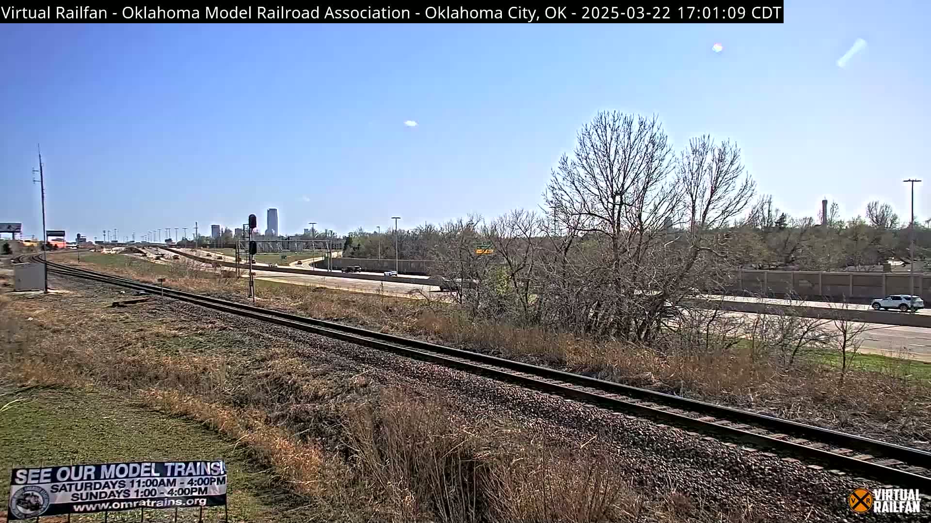 A sunny day reveals railroad tracks alongside a highway, with sparse, leafless trees and a distant cityscape under a clear blue sky.