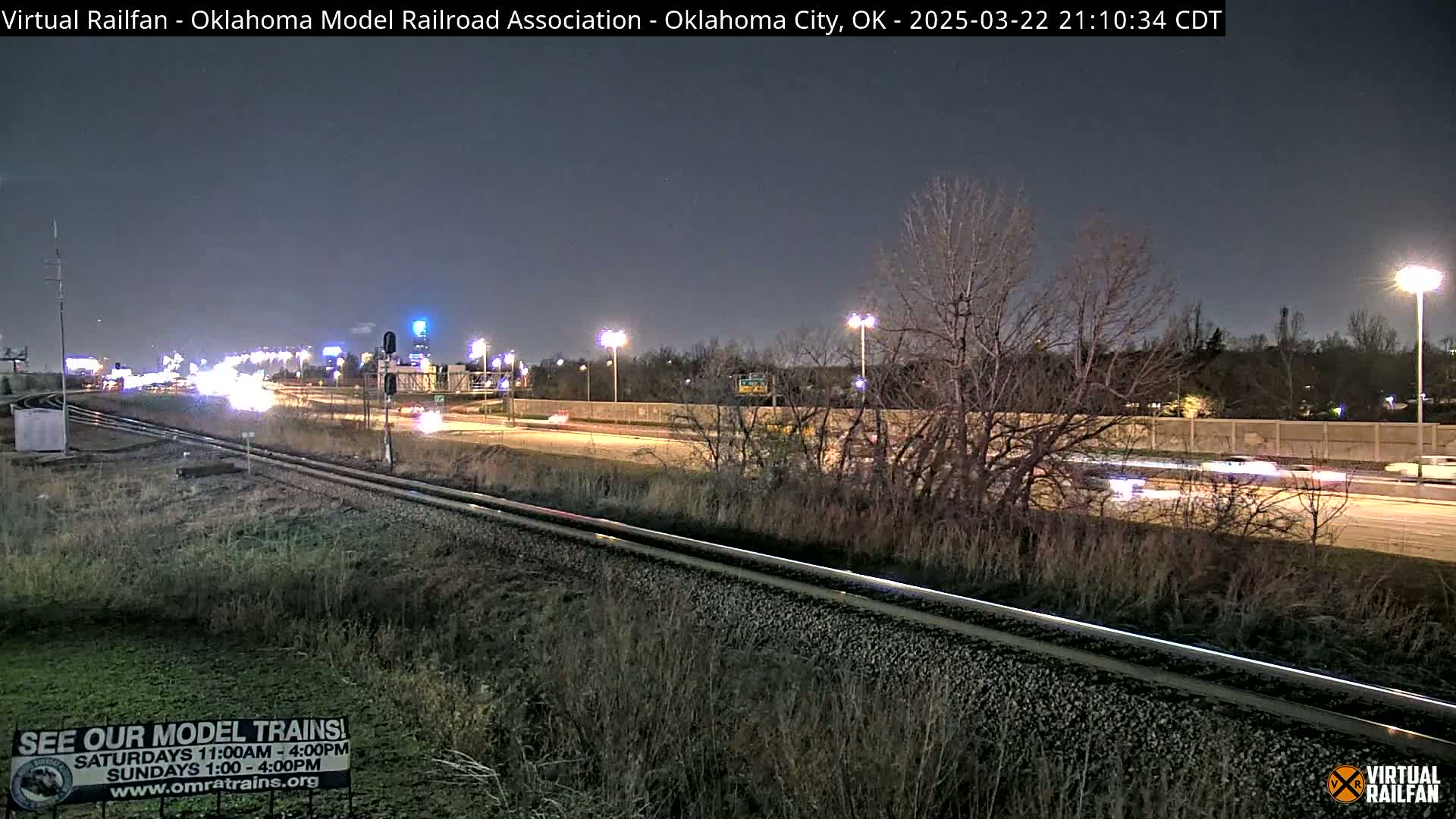 A nighttime view of a highway and parallel railroad tracks, with sparse traffic visible under a clear, dark sky.