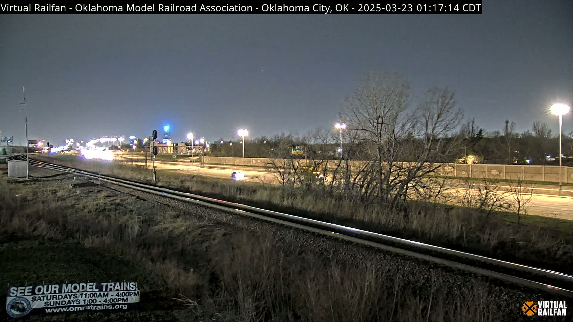 A nighttime view of railroad tracks beside a highway with sparse, leafless trees and a few cars visible under a dark sky.
