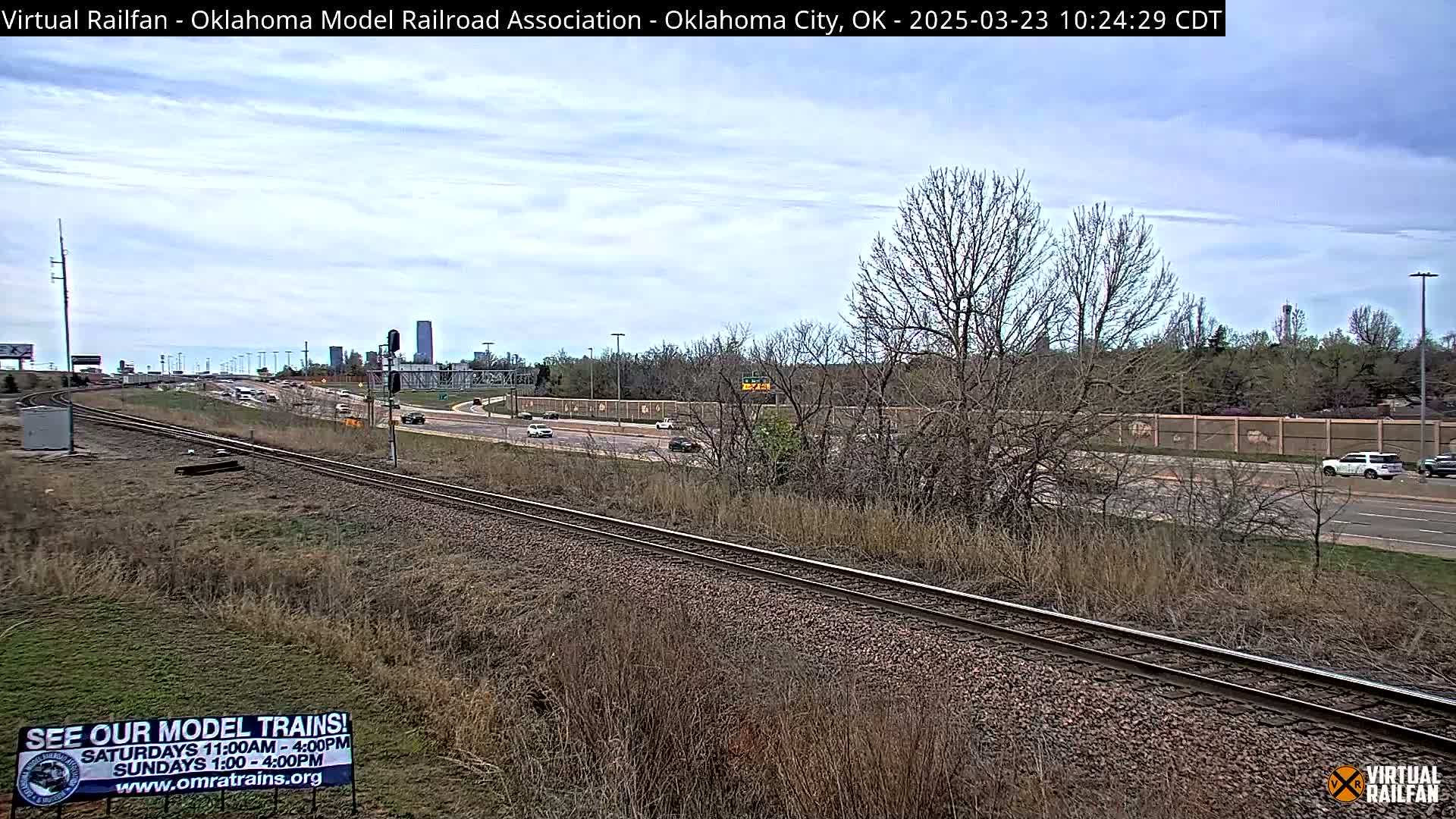 A partly cloudy day reveals a railroad track running alongside a highway with sparse, dry vegetation and a few cars visible on the road, and buildings in the distance.