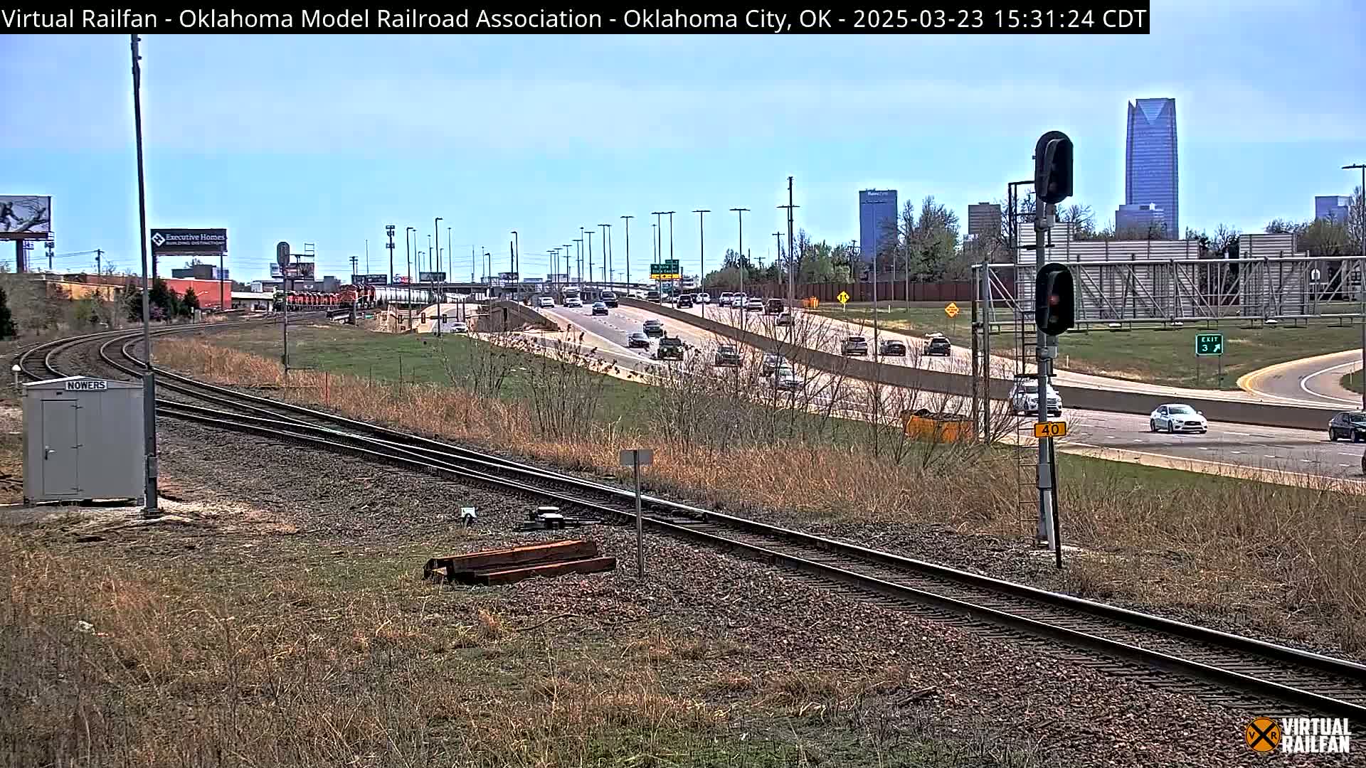 Railroad tracks curve alongside a highway with moderate traffic under a clear sky, with a cityscape including tall buildings visible in the background.