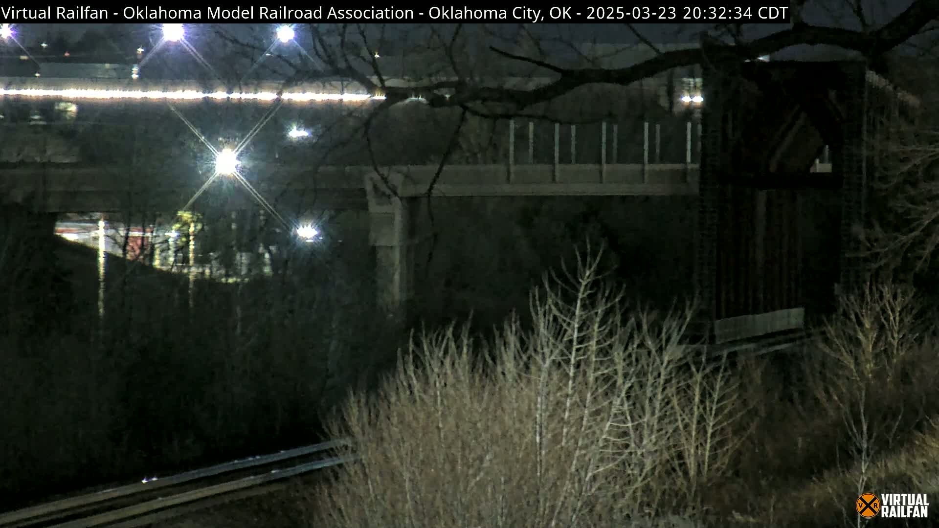 At night, under a dark sky, a view from a low vantage point shows a roadway overpass and railroad tracks, with bare winter shrubs in the foreground.