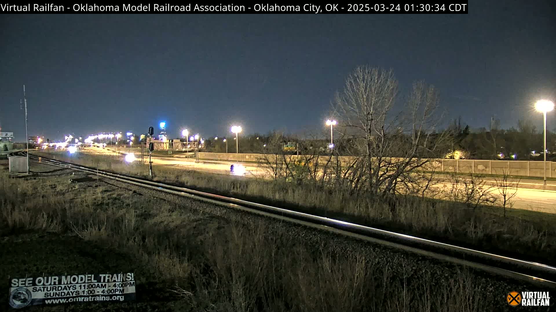 A nighttime view of railroad tracks beside a highway with sparse, leafless trees and low vegetation under artificial lighting.