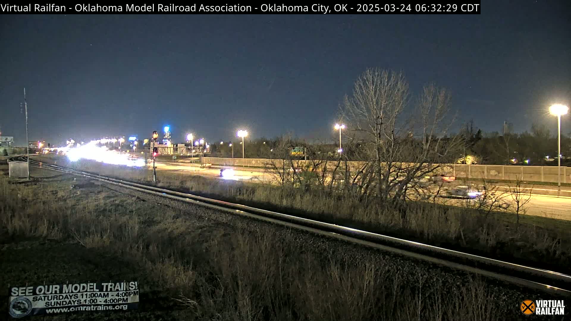 A nighttime view of a highway with sparse traffic, alongside a set of train tracks, under a clear, dark sky.