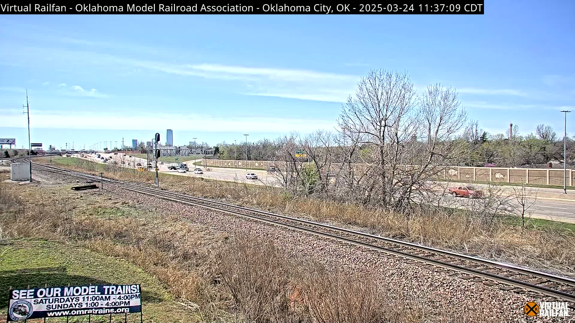 A partly cloudy sky overlooks a highway and train tracks, with sparse, dry vegetation alongside the tracks and a few cars visible on the road.