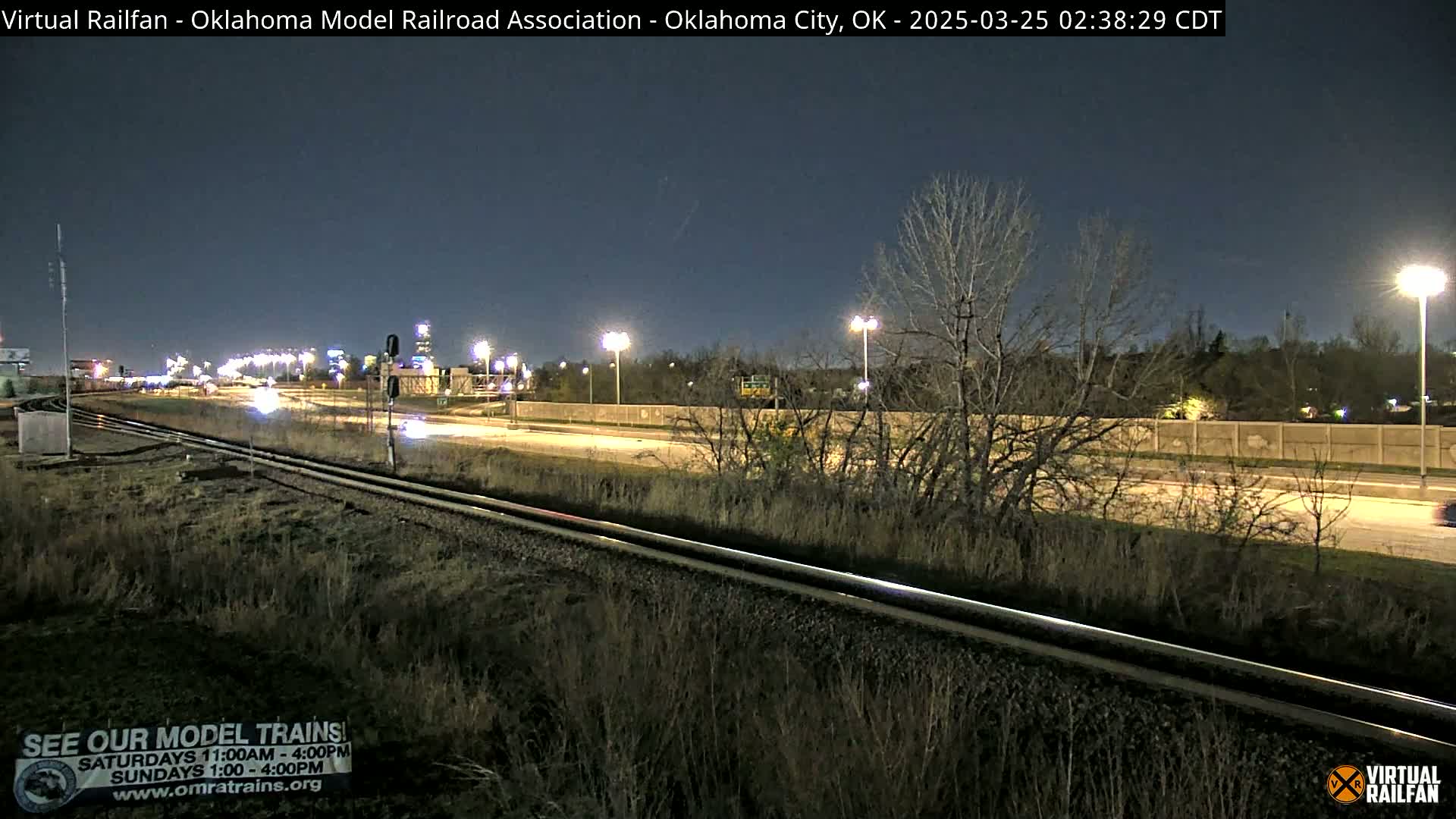 A nighttime view of a highway and railway tracks, with sparse vegetation and bare trees under a dark sky.