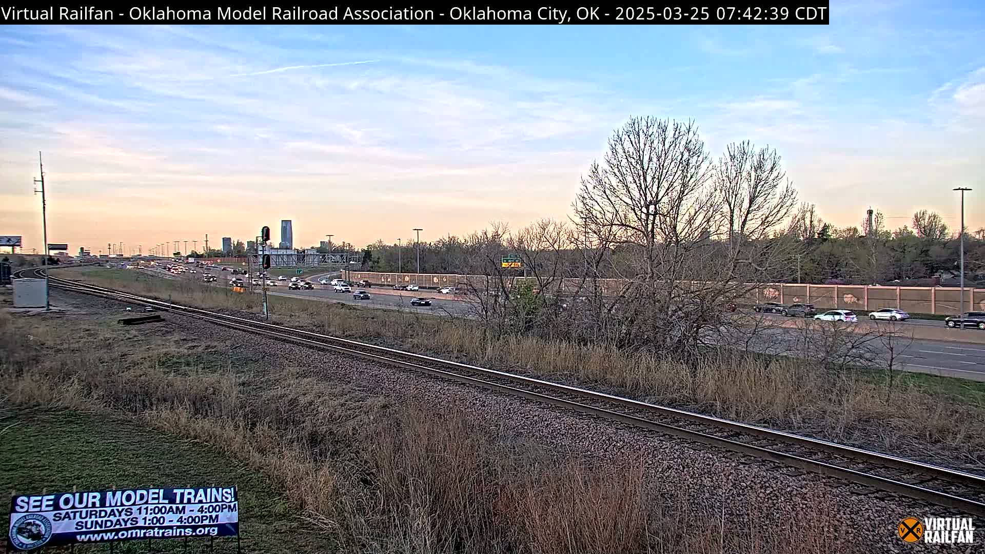 A railroad track runs alongside a highway with light traffic under a partly cloudy, pale-orange sunset sky.