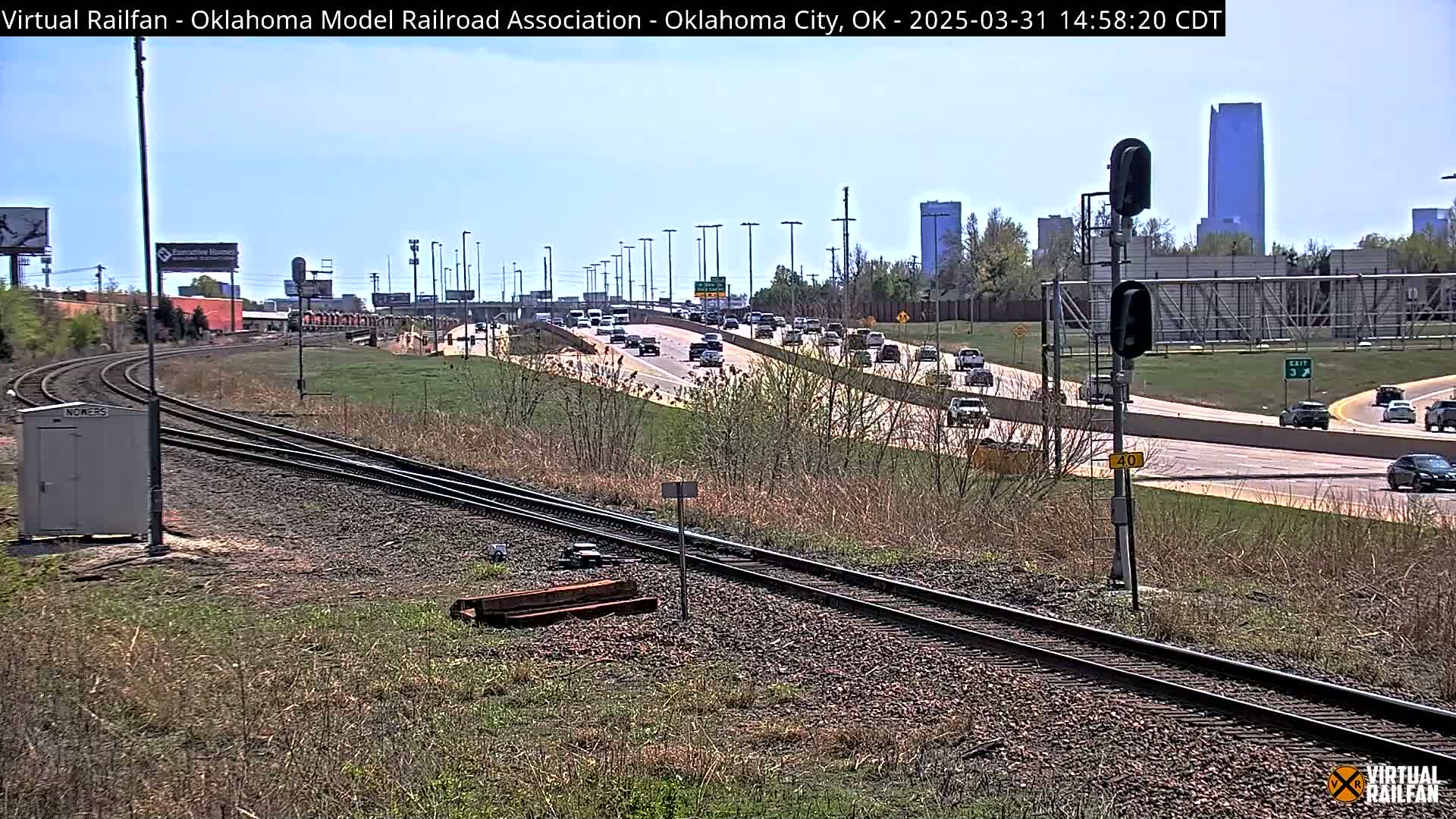 A sunny day shows a view of train tracks next to a busy highway, with a city skyline in the background.