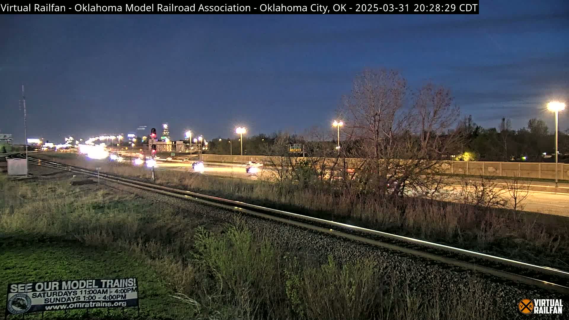 A nighttime view of a highway and railroad tracks, with sparse traffic and bare trees under a dark blue sky.