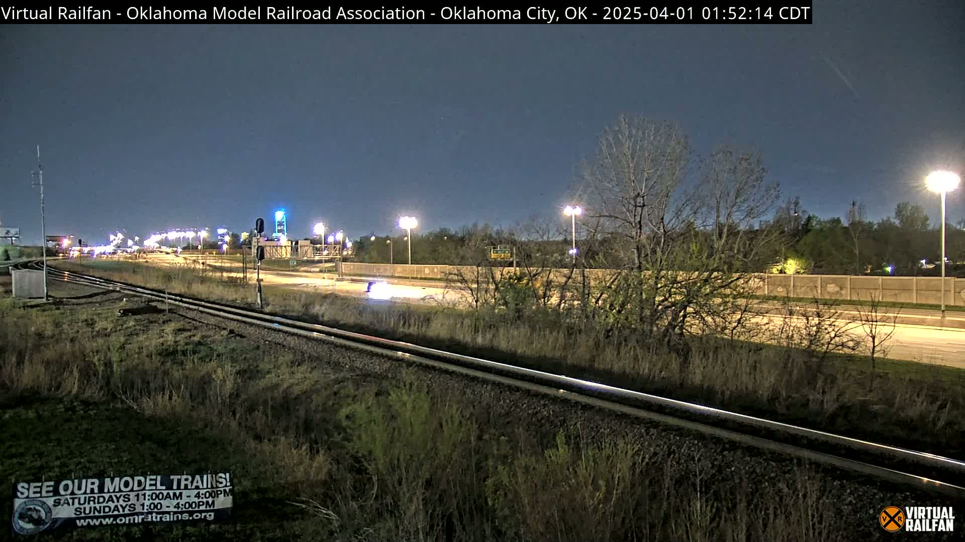 A nighttime view of railroad tracks next to a highway, under a clear, dark sky, with sparse vegetation along the sides.