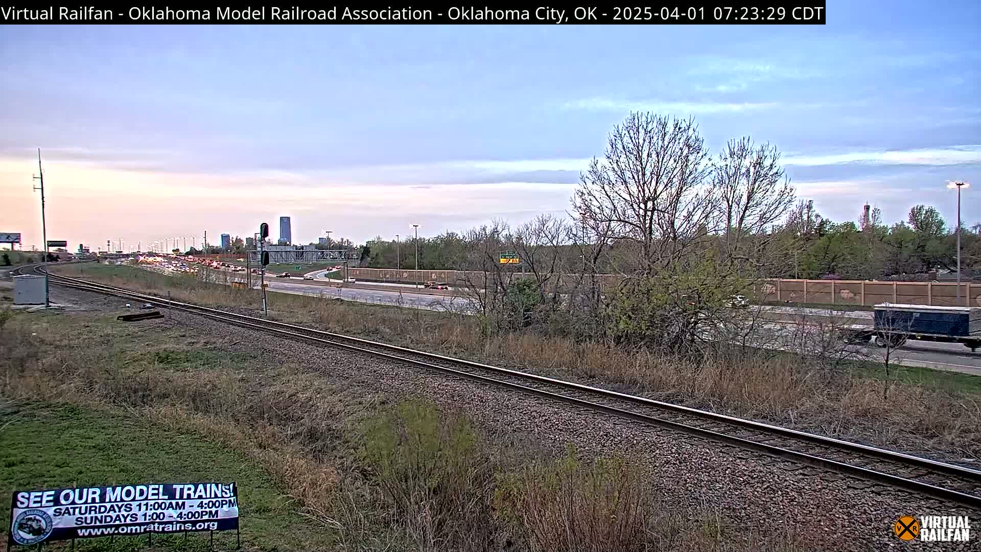 A railroad track runs alongside a highway with heavy traffic under a partly cloudy twilight sky.