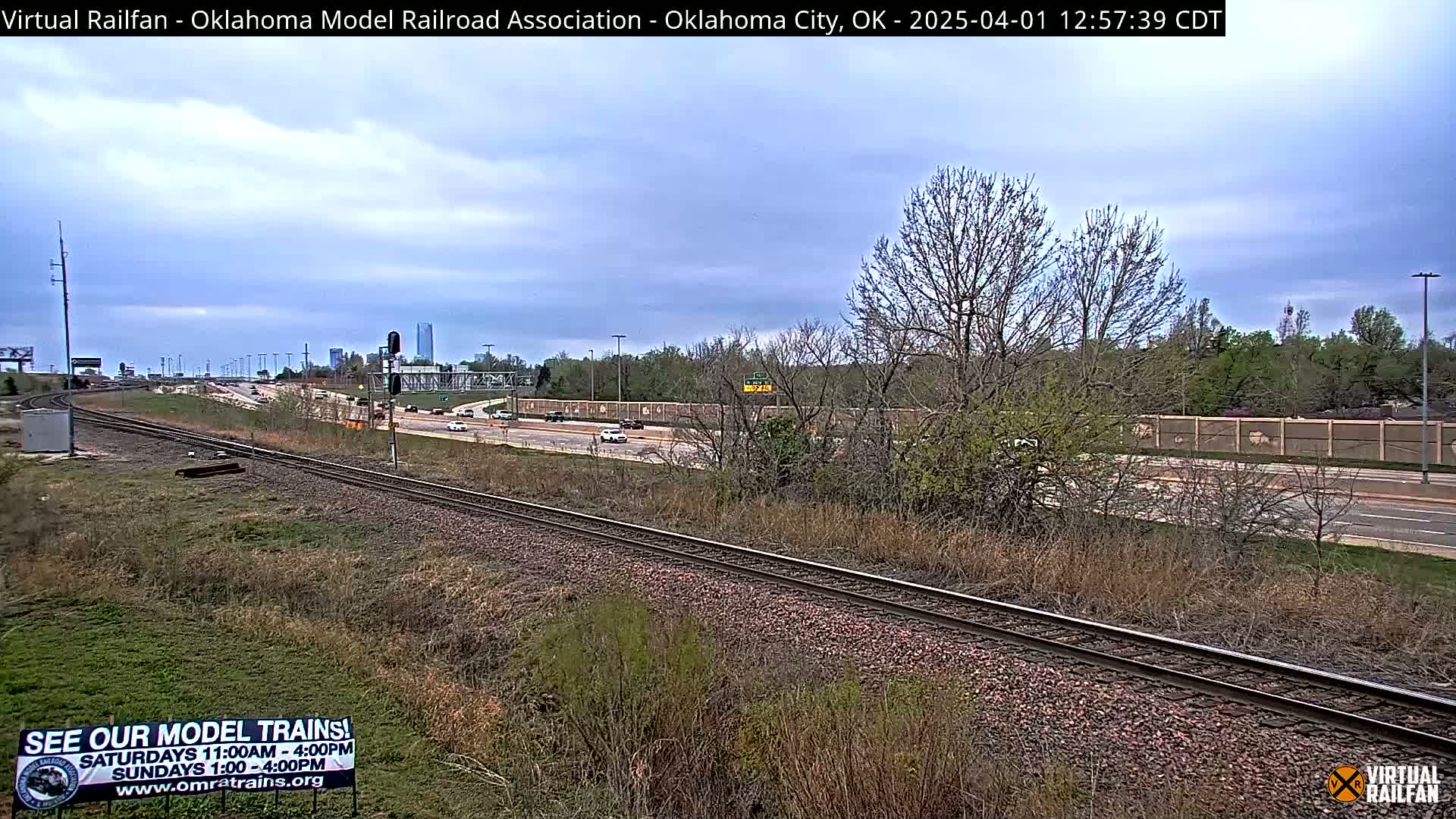 A mostly cloudy day reveals railroad tracks next to a highway with sparse traffic, passing by a low wall and some trees.