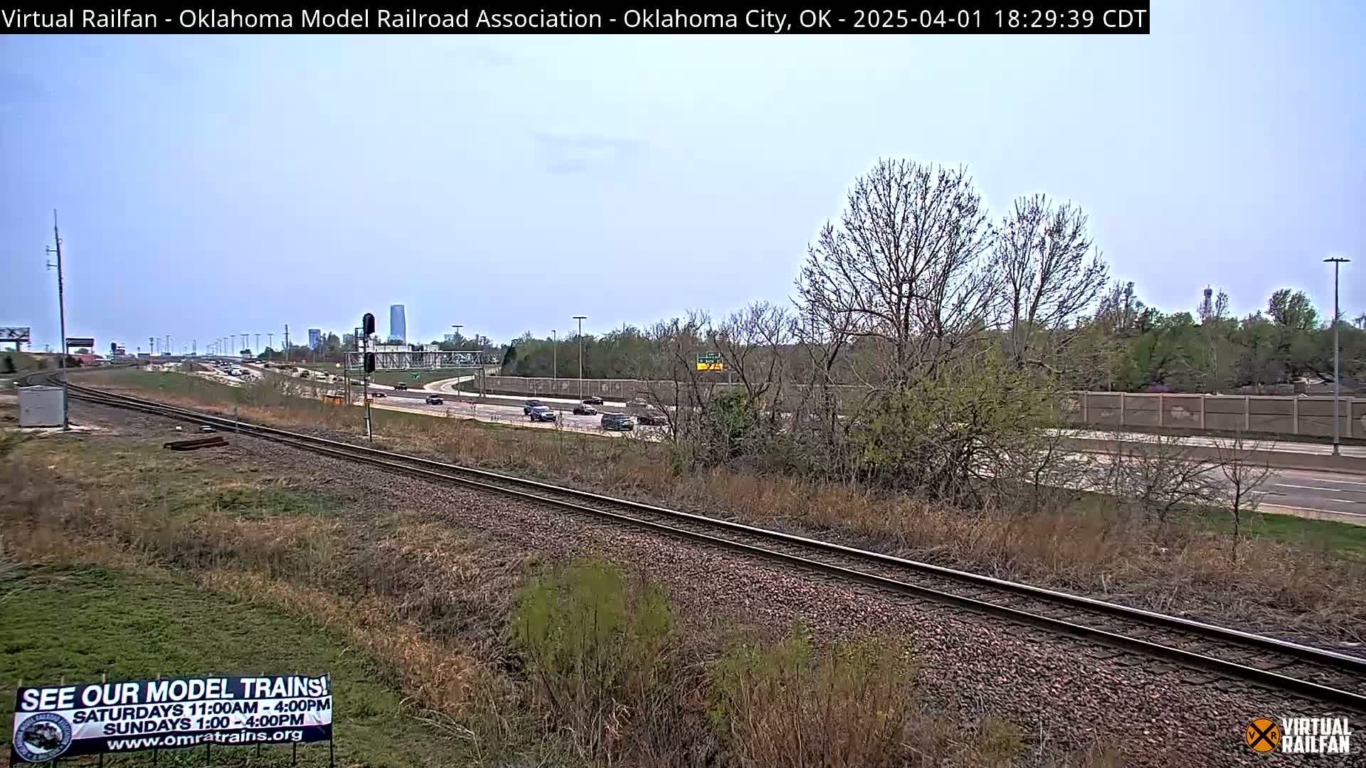 A mostly clear, daytime view of railroad tracks alongside a highway, with sparse early spring vegetation and distant buildings visible.