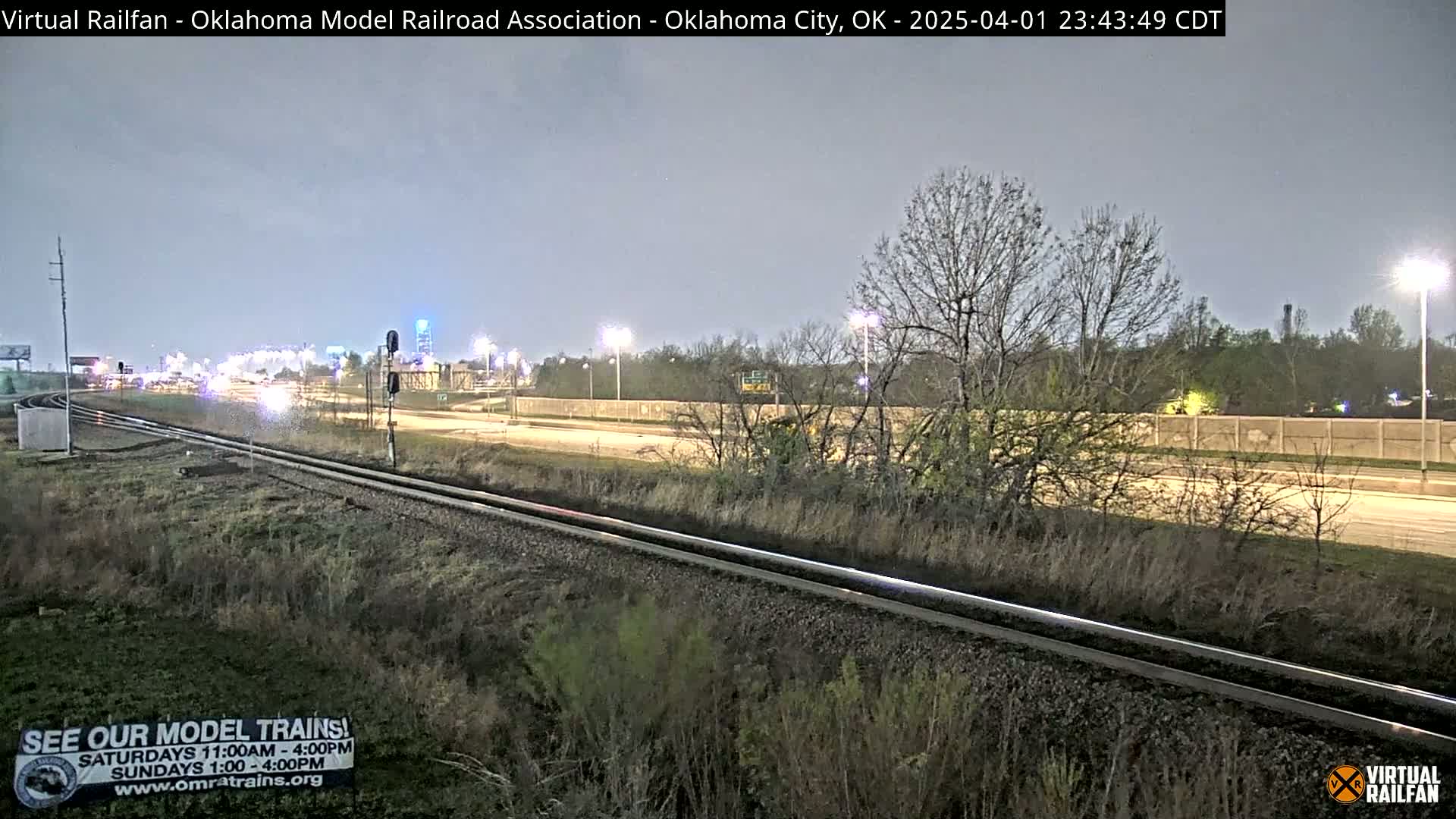 A nighttime view of railroad tracks running parallel to a highway, with bare trees and sparse vegetation along the sides.