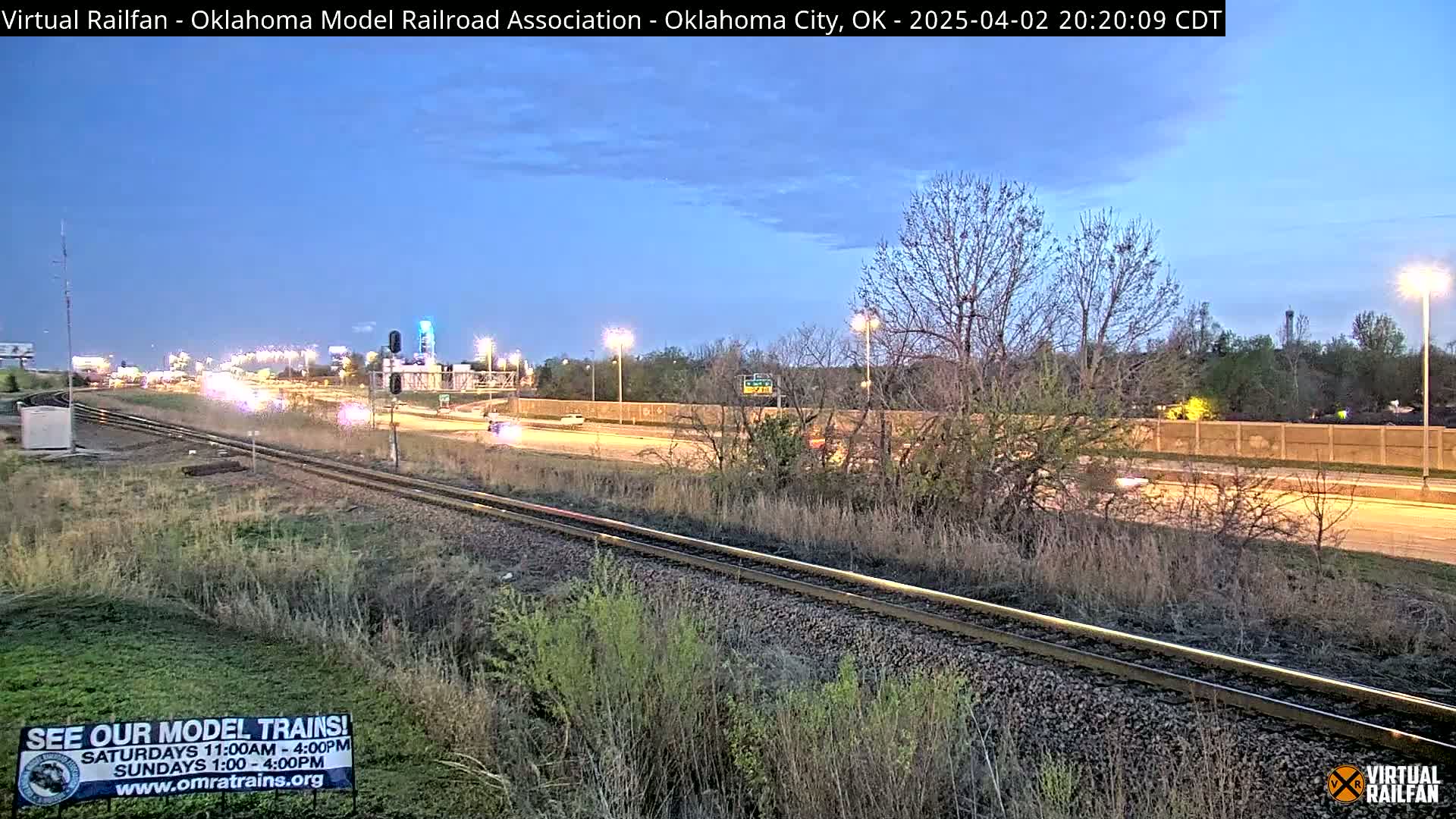 A twilight view shows train tracks next to a highway with sparse traffic under a clear, dusky sky.